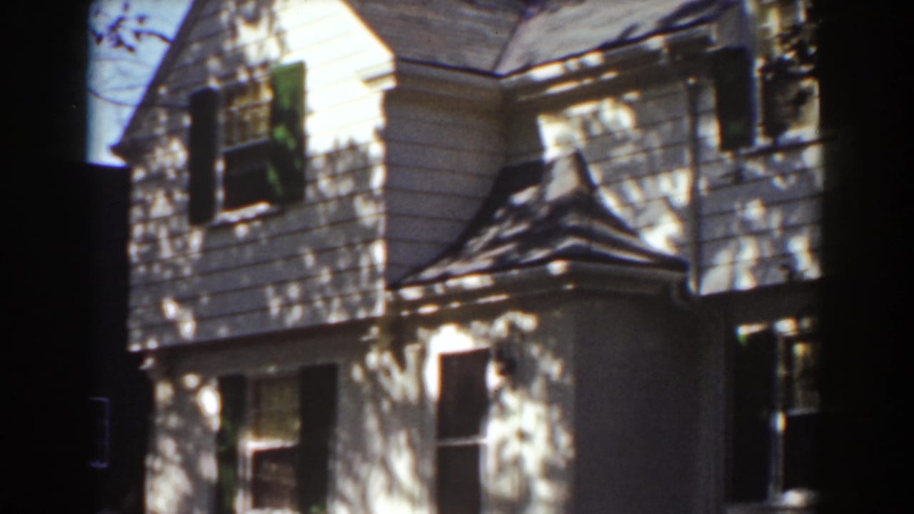 White clapboard house with dark shutters seen through tree shade.