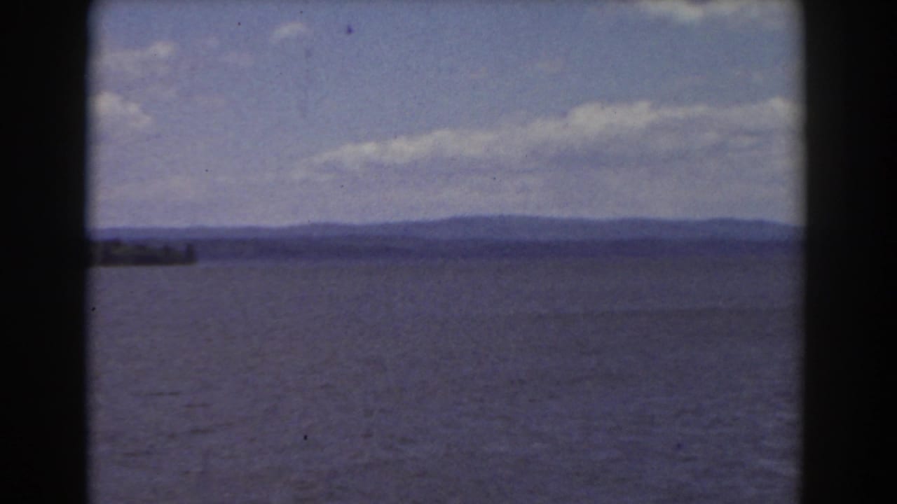 Wide lake with rippled water, distant shoreline, hazy hills, and partly cloudy sky.