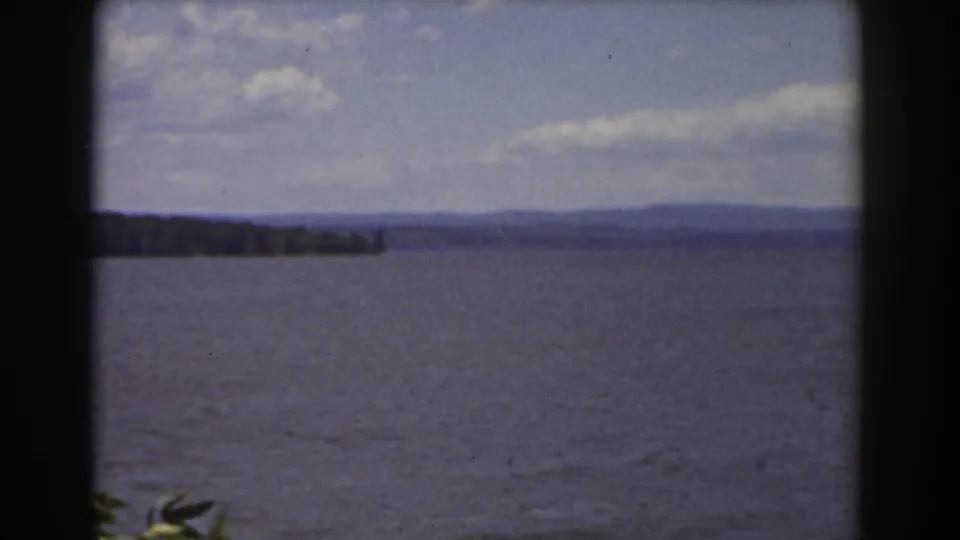 1939 New Hampshire lake view with open water, a distant wooded point, hazy hills, scattered clouds, and visible 8mm frame edges.