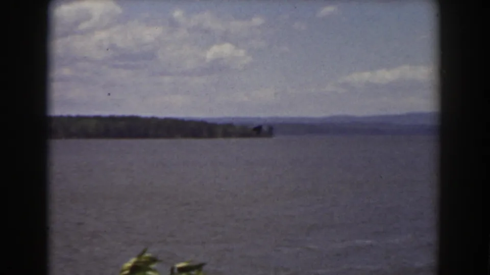 Home-movie frame from the Mount Washington, New Hampshire metadata showing lake water, a long tree-lined shore, low hills, clouds, and foreground leaves.
