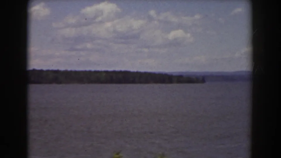 1939 8mm lake scenery near Mount Washington, showing rippled water, a wooded shoreline, hazy hills, partly cloudy sky, and dark rounded frame edges.