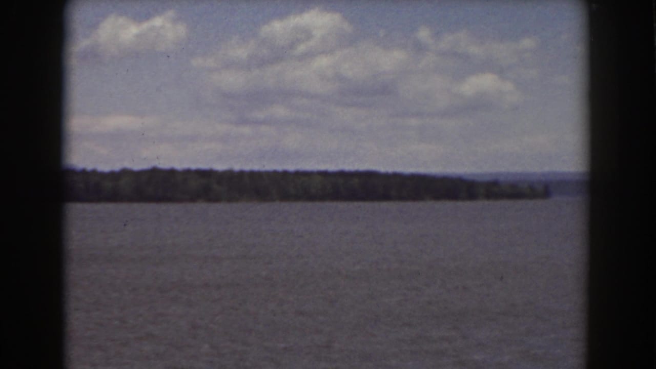 Wide water view with distant tree-lined shore, low mountains, clouds, and dark 8mm film borders.