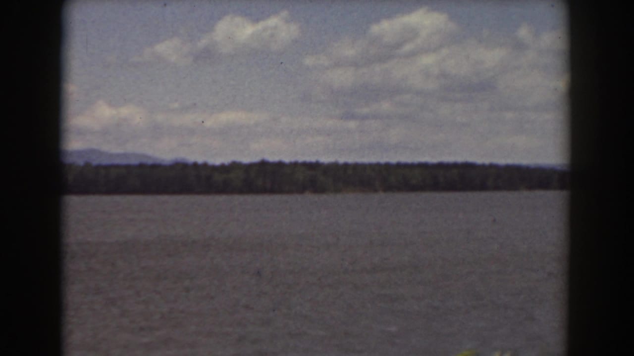 Lake or river surface with a dark wooded shoreline, distant hills, clouds, and visible film gate edges.