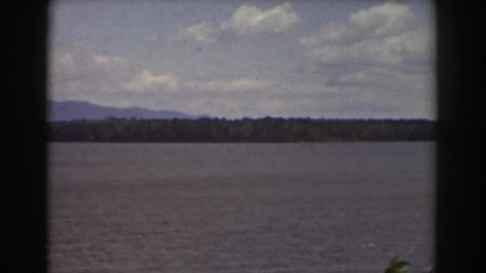 1939 New Hampshire home-movie frame of a calm waterway facing a dark shoreline and low mountains under scattered clouds.