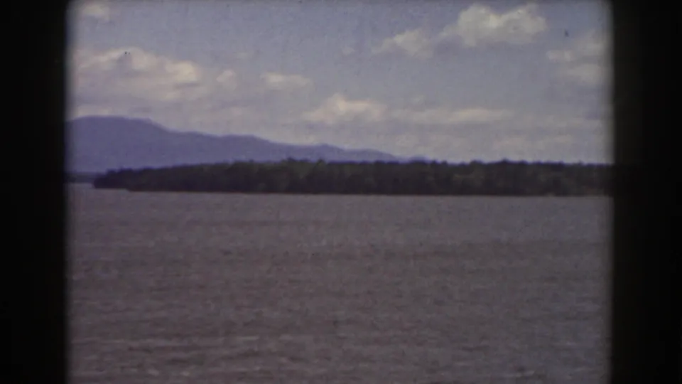 Mount Washington-area 8mm landscape showing open water, a wooded shoreline, and blue-gray mountains beneath a partly cloudy sky.