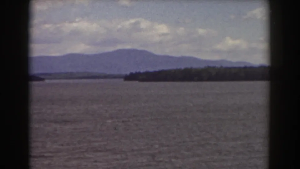 Vintage 8mm frame of a broad water surface, low wooded shoreline, and distant New Hampshire mountains beneath scattered clouds.