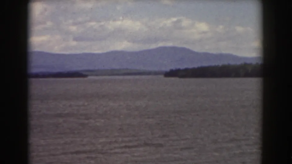1939 home-movie landscape view showing blue-gray water, hazy mountains, low clouds, and a wooded point along the shore.