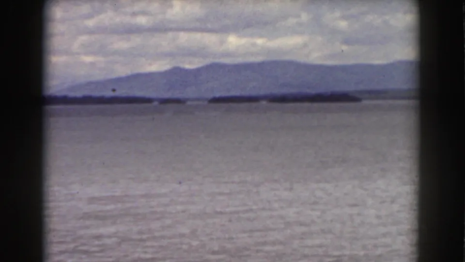 1939 New Hampshire landscape frame with calm water, a low wooded island, and hazy mountain slopes in the distance.