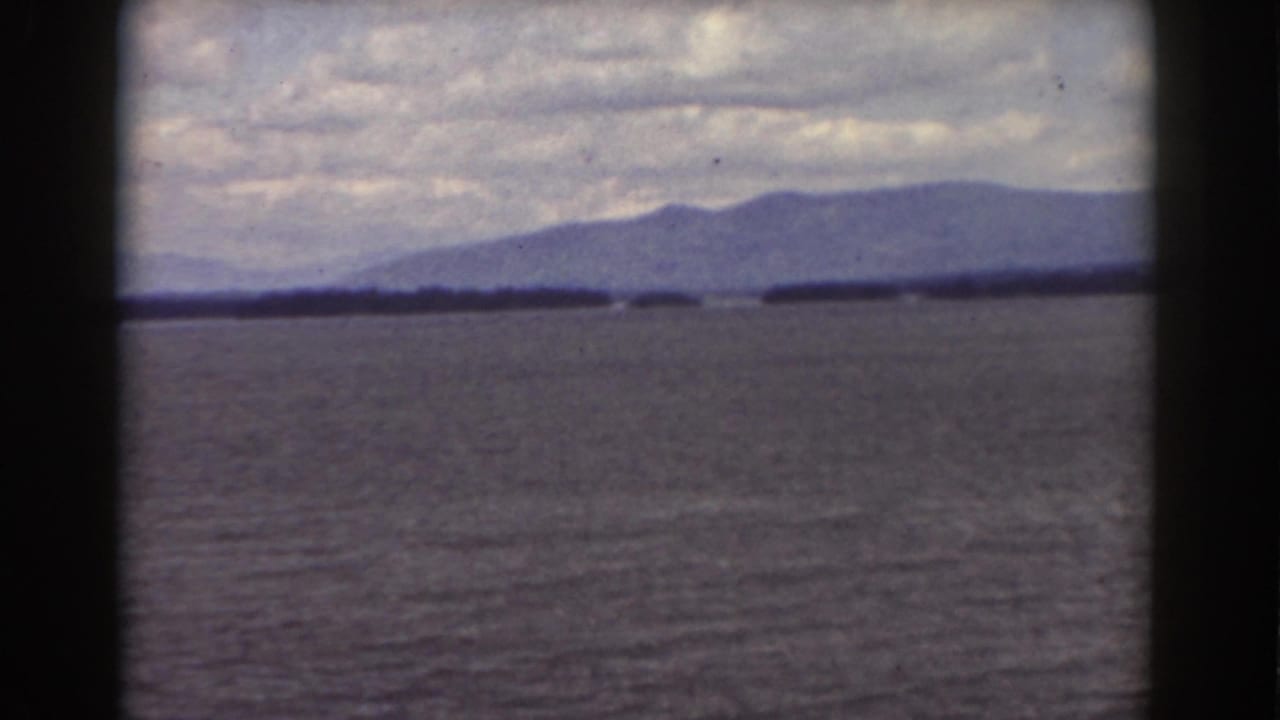 Calm rippled water with a low shoreline and distant hazy mountains under a cloudy sky.