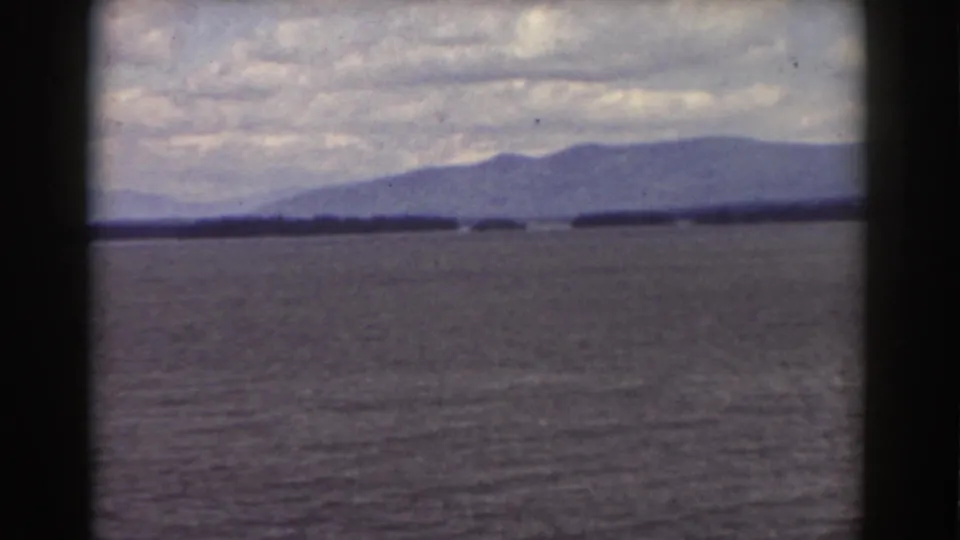 1939 8mm Mount Washington-area view of calm water, a low dark shoreline, and distant hazy mountains beneath cloud cover.