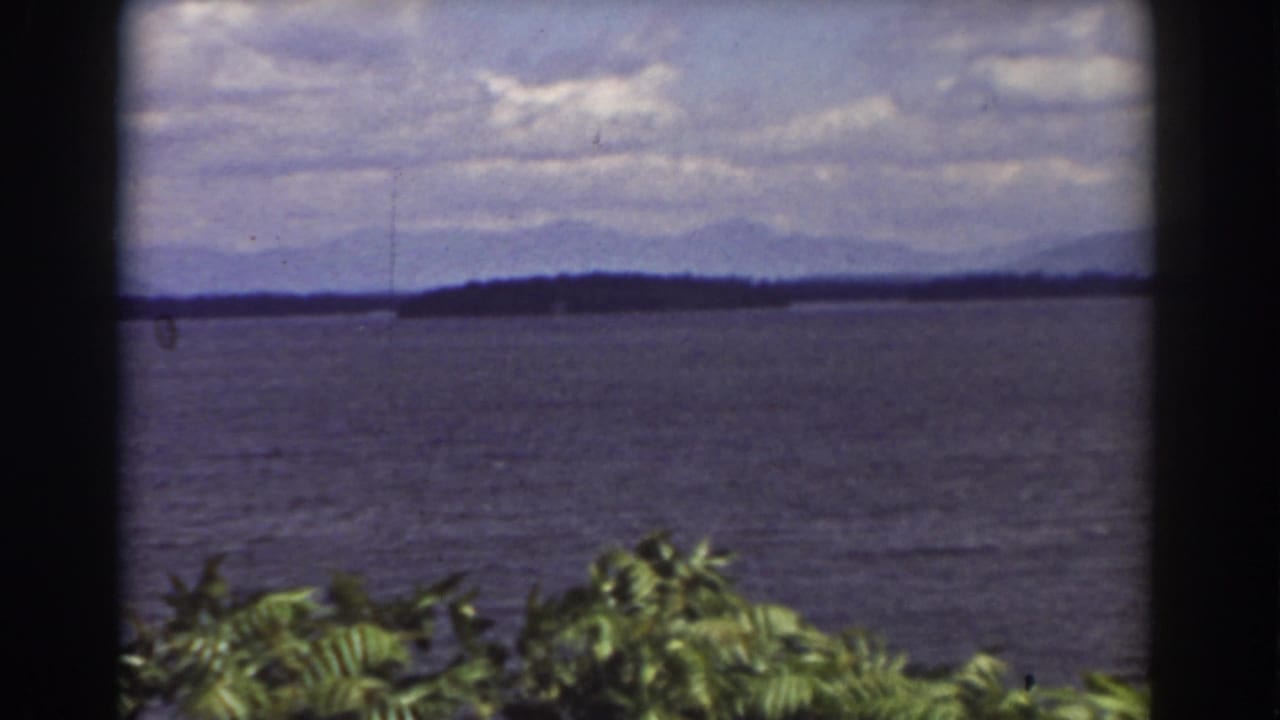 1939 lakeside 8mm frame near Mount Washington with open water, a low shoreline, blue mountain silhouettes, clouds, and windblown foreground plants.