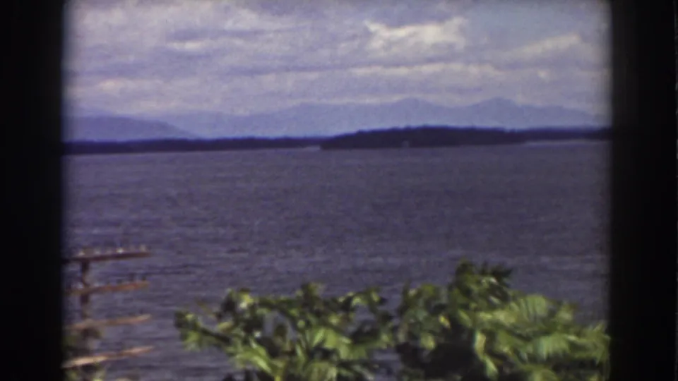 1939 Mount Washington-area lake view with leafy foreground plants, a small dock or railing at left, open water, distant wooded hills, and clouds.
