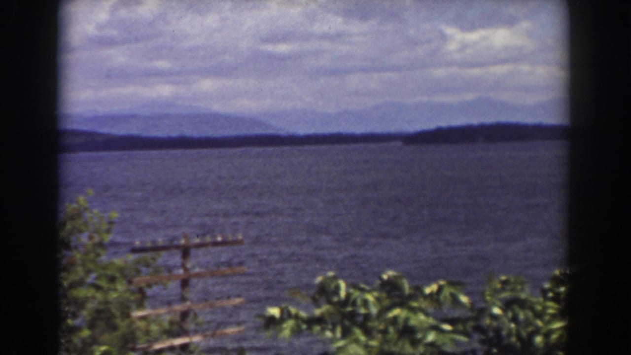 1939 8mm lakeside frame near Mount Washington showing windblown leaves, a small railing or dock, broad water, distant shoreline, and hazy mountains.