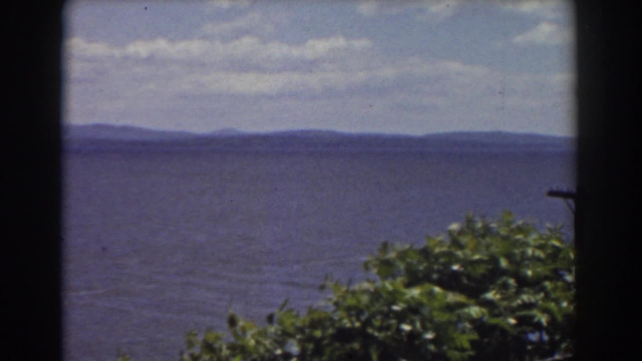 1939 New Hampshire home-movie frame showing water, mountain ridges, broken clouds, and dense green foliage in front.