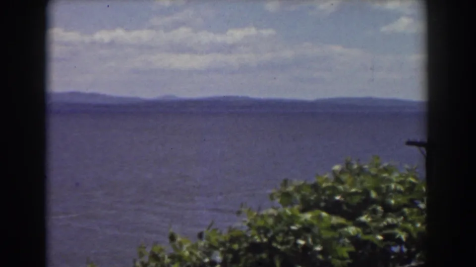 1939 New Hampshire home-movie frame showing water, mountain ridges, broken clouds, and dense green foliage in front.