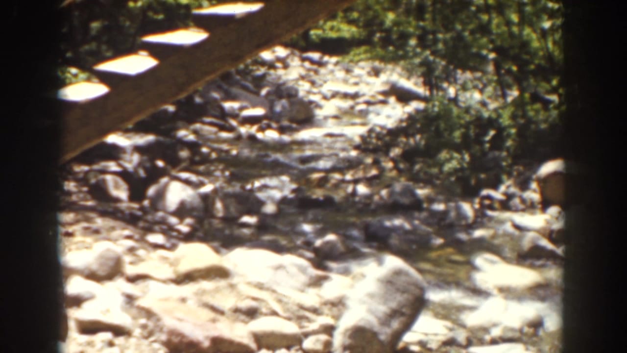 Rocky wooded stream in the Mount Washington, New Hampshire clip, viewed from under a wooden structure with sunlight on stones and water.