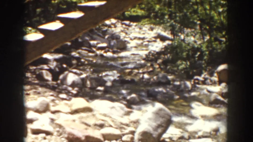 Rocky wooded stream in the Mount Washington, New Hampshire clip, viewed from under a wooden structure with sunlight on stones and water.
