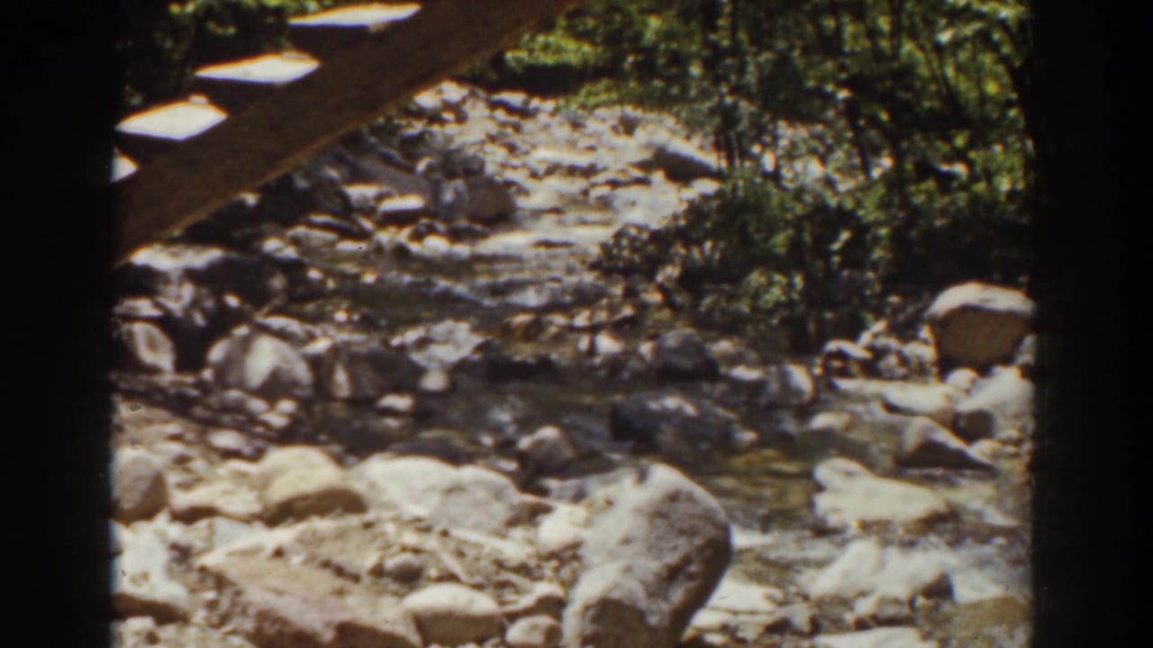 1939 New Hampshire home-movie frame of a shallow rocky stream and wooded bank, partly covered by a wooden overhead structure.