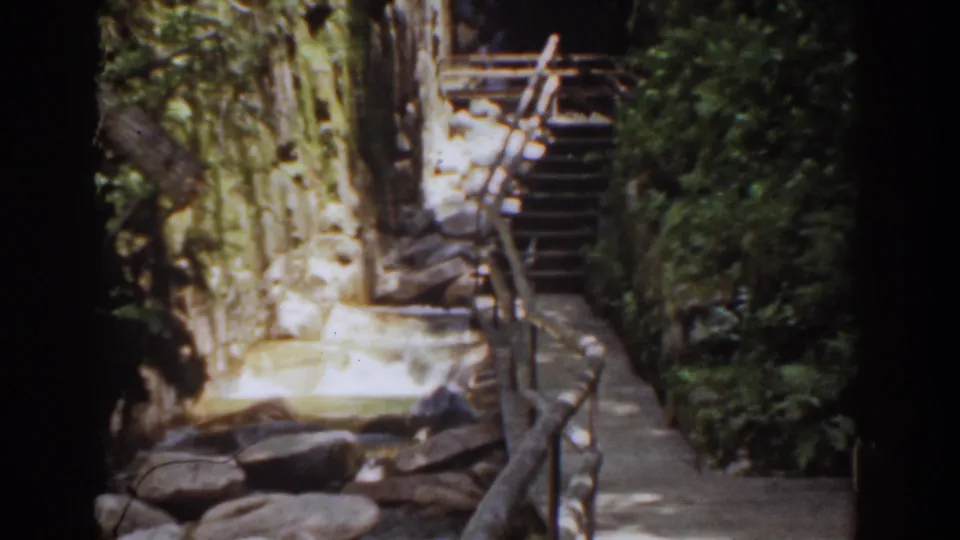 1939 Mount Washington home-movie frame showing a stone path, rustic railing, rock walls, trees, and steps through a shaded mountain setting.