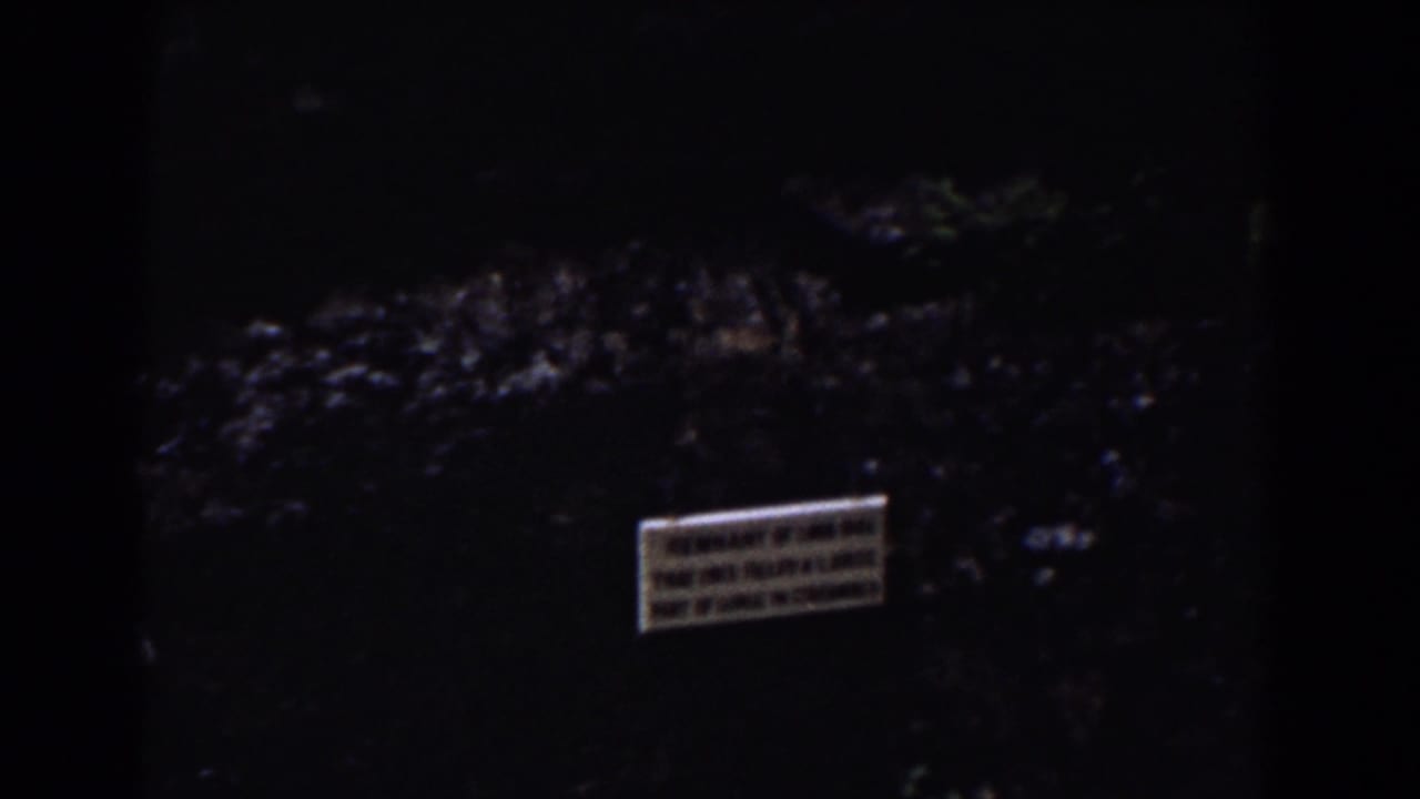 1939 Mount Washington frame with a small posted sign on a shadowed rock face beside dark water and surrounding vegetation.