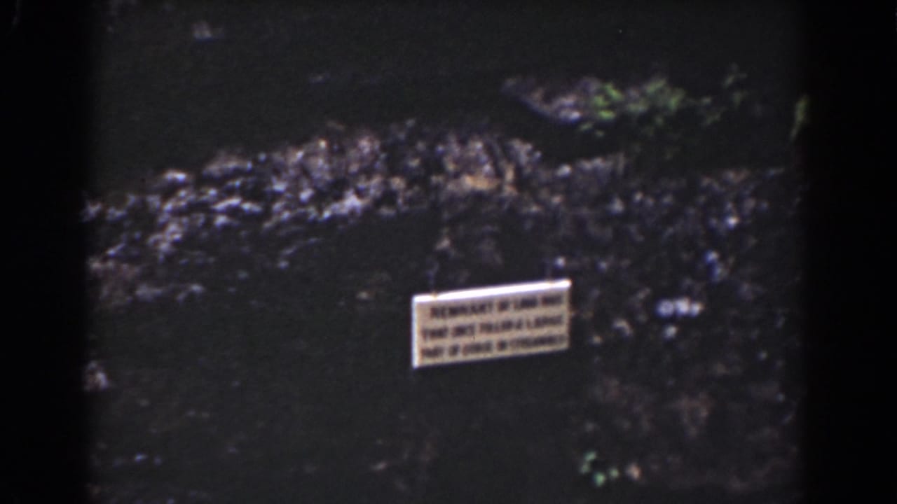 1939 Mount Washington home-movie frame showing a small sign near dark water, rocky edges, and patches of green vegetation.