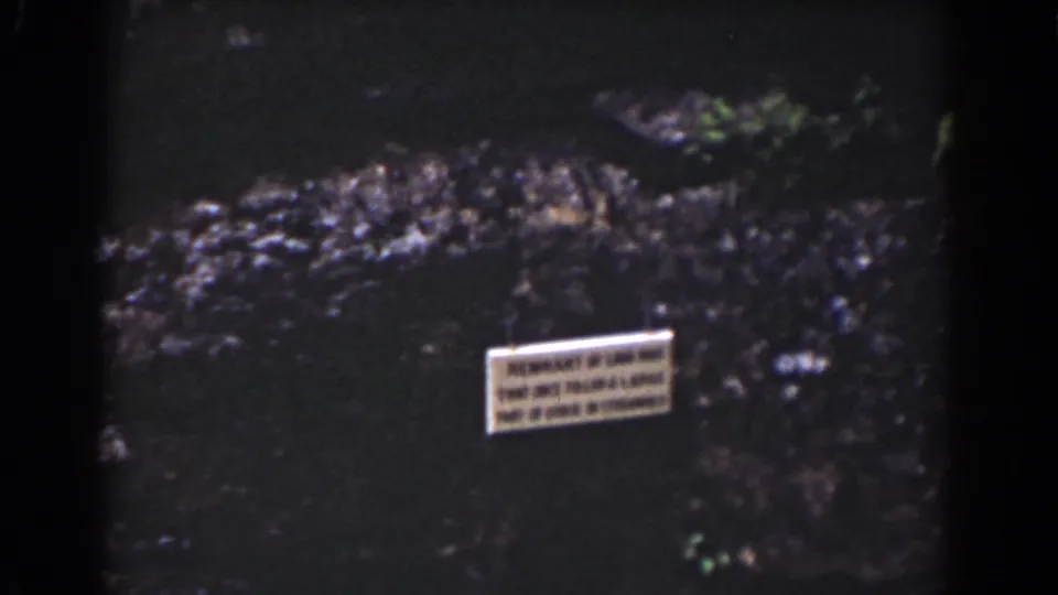 1939 Mount Washington home-movie frame showing a small sign near dark water, rocky edges, and patches of green vegetation.