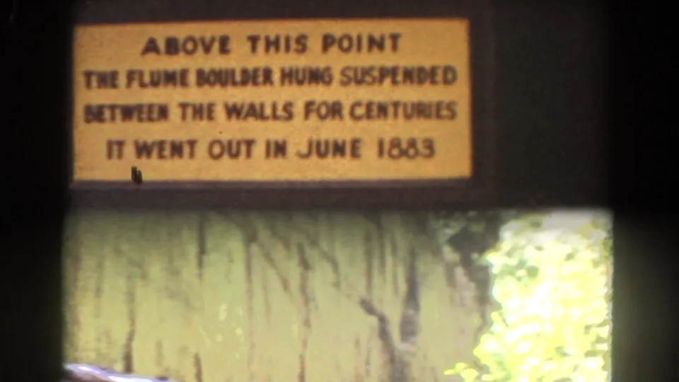 The camera frames the Flume Boulder plaque with rough stonework and sunlit foliage below in New Hampshire travel footage.