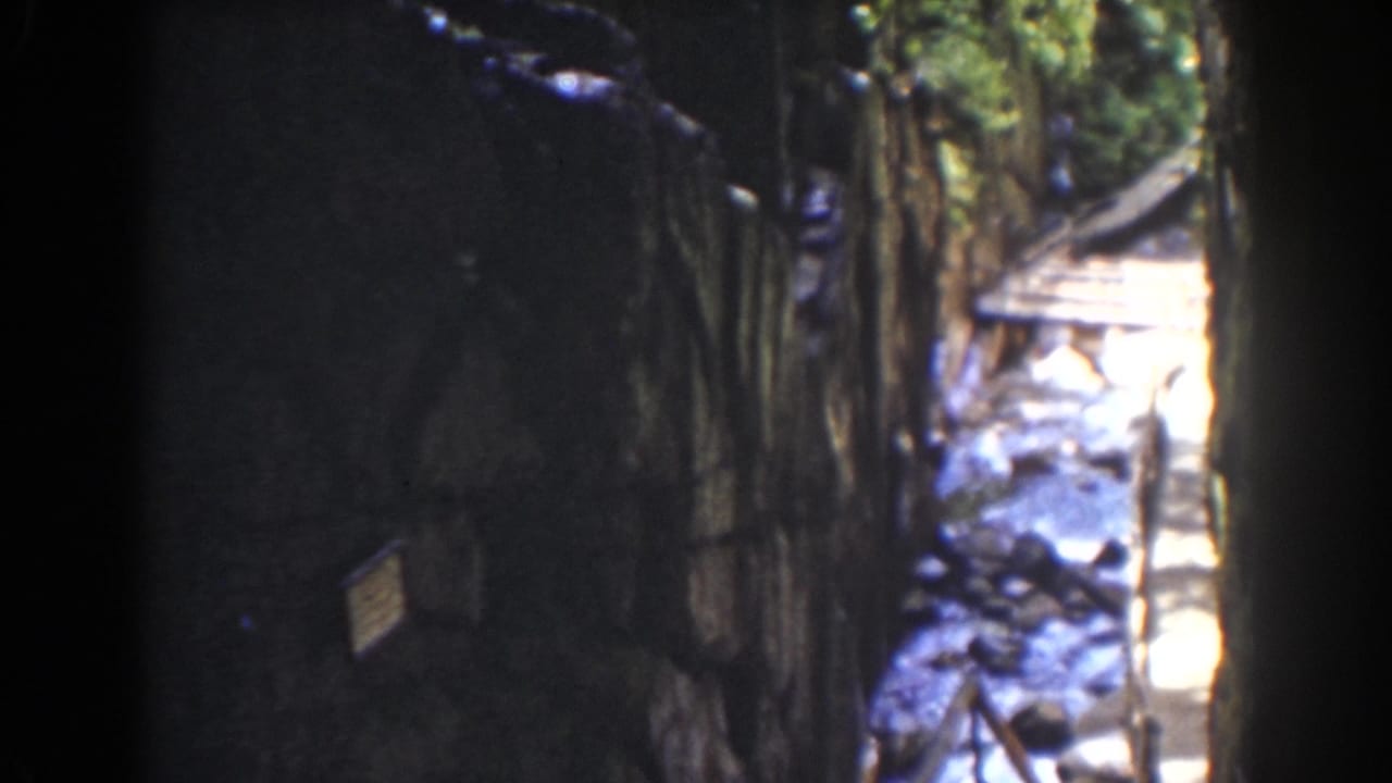 1939 Mount Washington frame with a dark rock or timber wall, a small sign-like pale patch, and bright water beside rough boards.