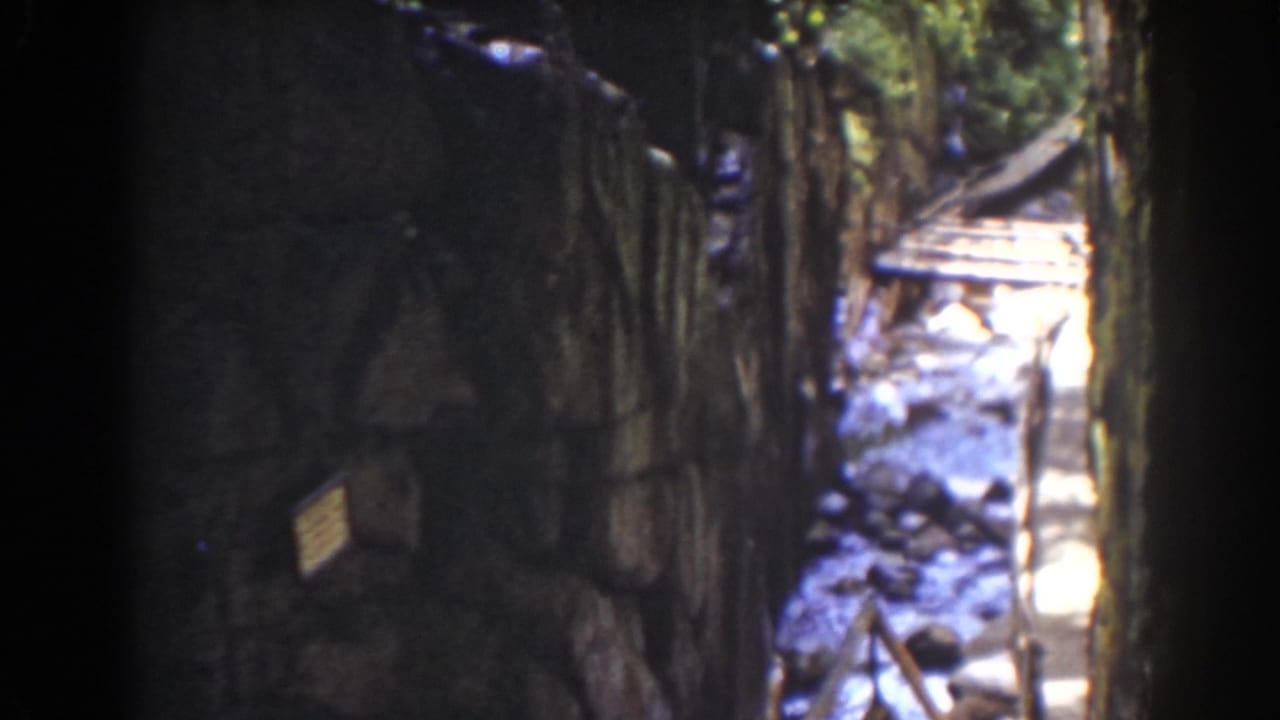 Dark stone-walled walkway beside bright rocky water and timber railings.