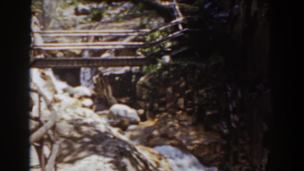 1939 Mount Washington home-movie frame showing a rustic wooden footbridge over rocks and flowing water in dense woods.
