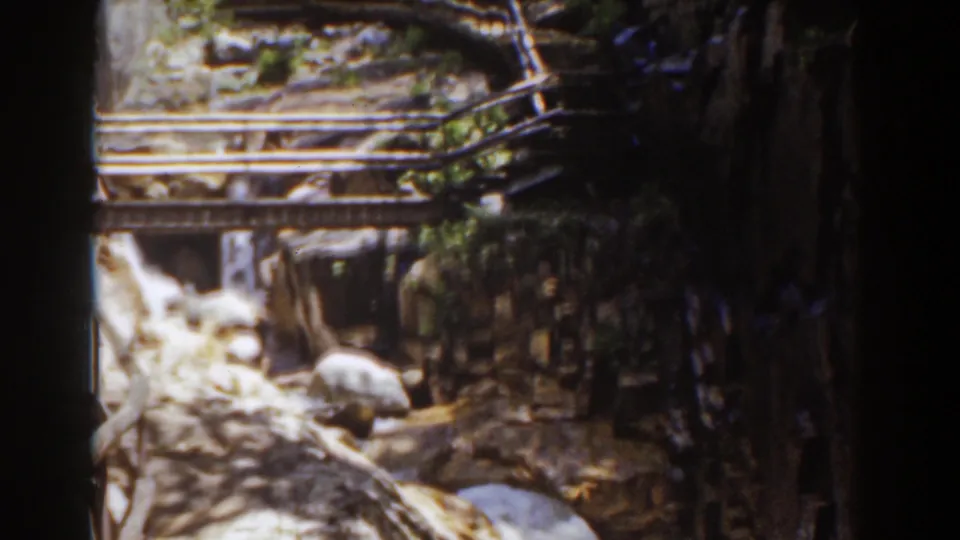 1939 Mount Washington 8mm frame with a wooden footbridge, boulders, and water running through a forested rocky channel.