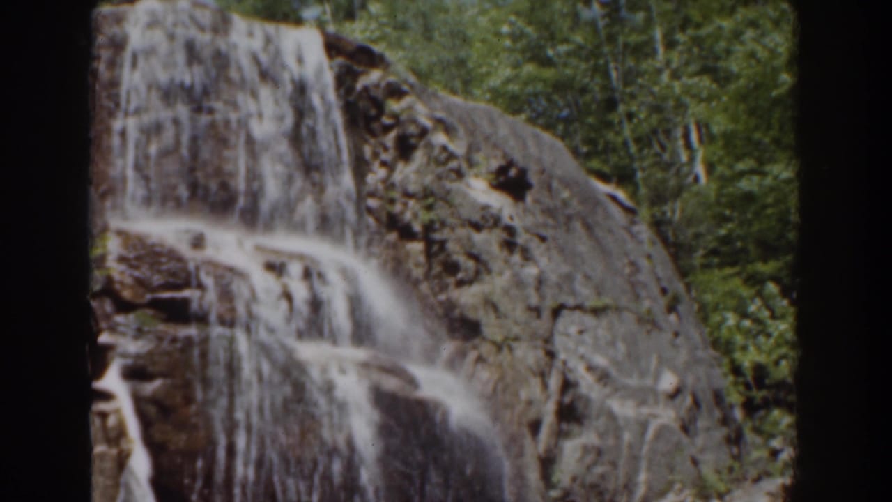 1939 Mount Washington home-movie frame showing water spilling over a brown rock face with forest in the background.