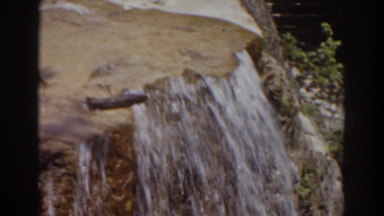 8mm Mount Washington frame showing water draining over a ledge, with a brown pool, branch-like debris, and plants near the rocks.