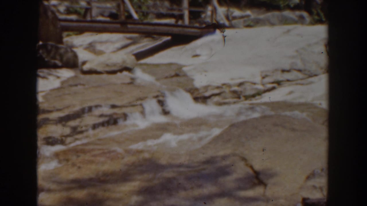 1939 home-movie frame near Mount Washington shows water moving over smooth rocks with a wooden footbridge or railing behind it.
