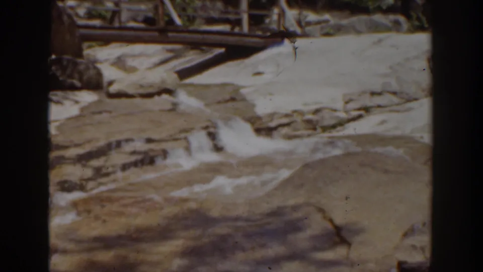 1939 home-movie frame near Mount Washington shows water moving over smooth rocks with a wooden footbridge or railing behind it.