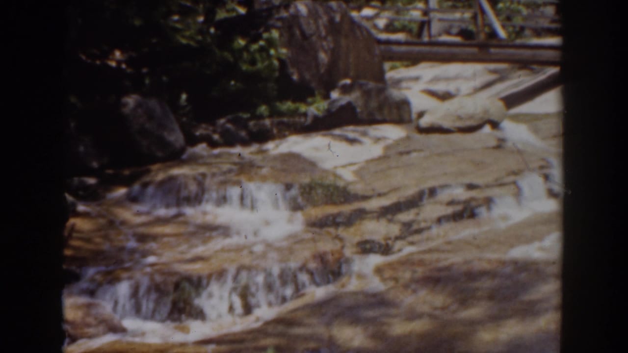 Mount Washington, New Hampshire home-movie frame of water crossing rocky ledges near a wooden bridge in bright daylight.