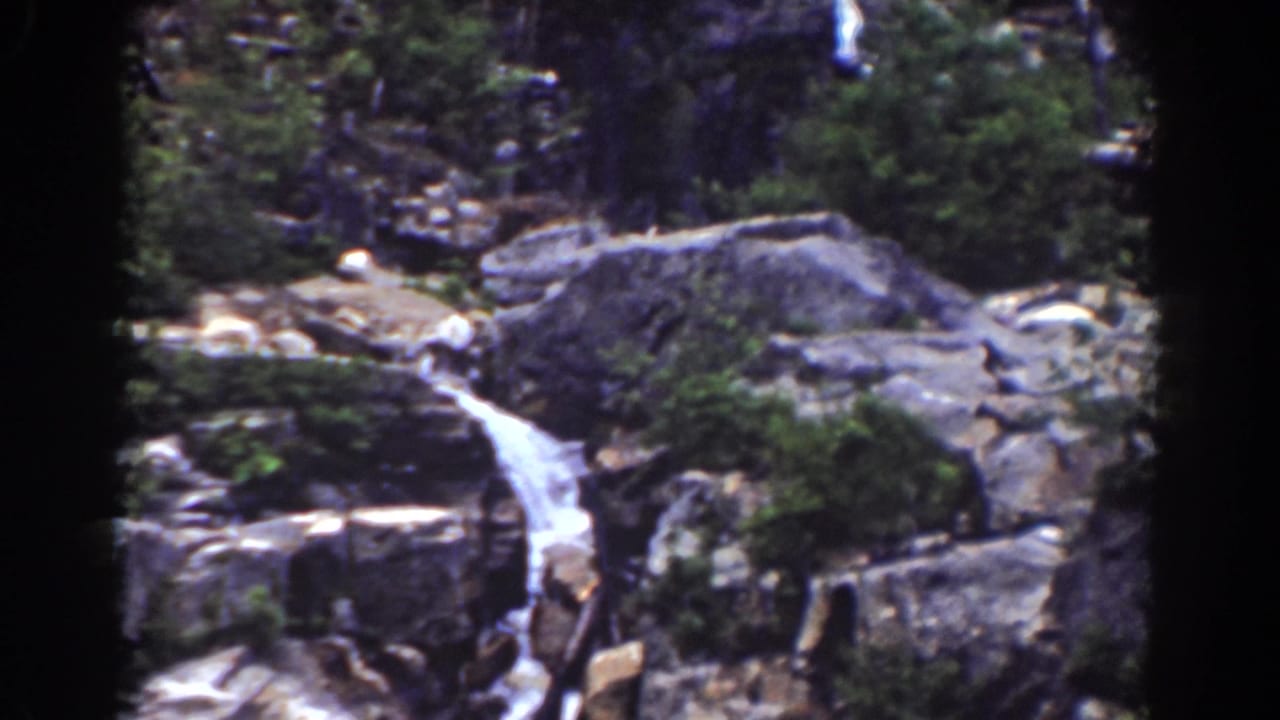 Small waterfall running over gray rock ledges with evergreen shrubs nearby.