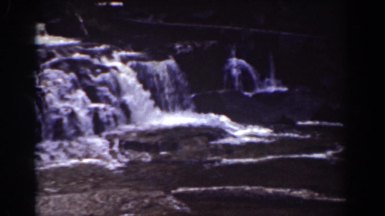 Waterfall spilling over layered rocks in a dark, grainy 8mm frame.