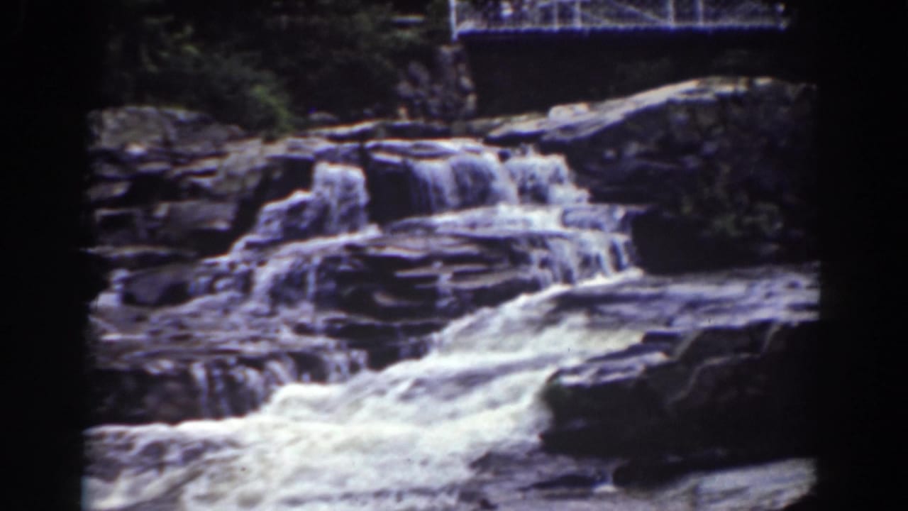 Water cascades over layered rocks below a white pedestrian bridge.