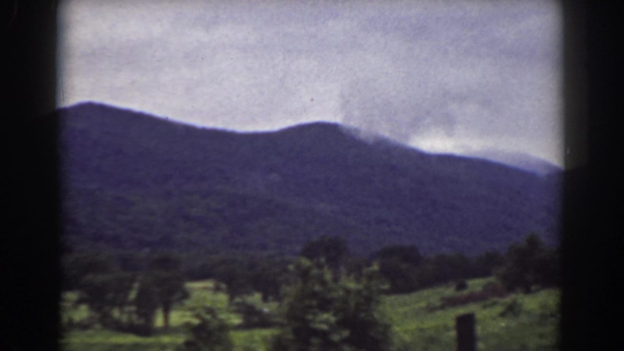 Vintage 8mm landscape frame from the Mount Washington area, showing a broad mountain ridge over trees and fields beneath an overcast sky.