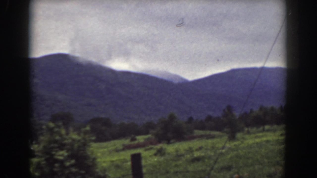 1939 8mm frame of the Mount Washington area, showing a green hillside, trees, fence post, and cloudy mountain backdrop.