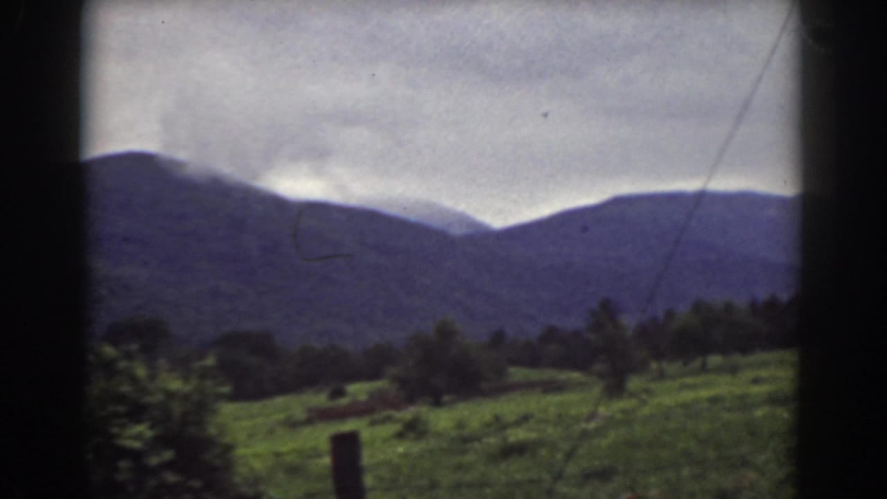 Green field and fence post below distant mountain ridges under a cloudy sky.
