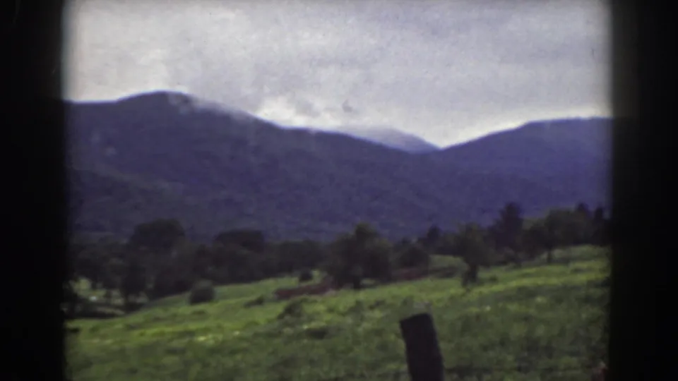 Archival 8mm view of rolling New Hampshire countryside near Mount Washington, with trees, a foreground post, and layered mountain slopes.