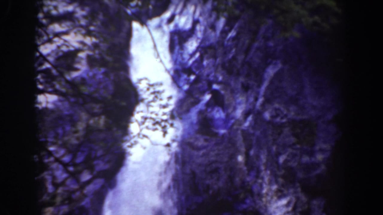 Waterfall dropping through a dark rocky gorge with sparse vegetation.