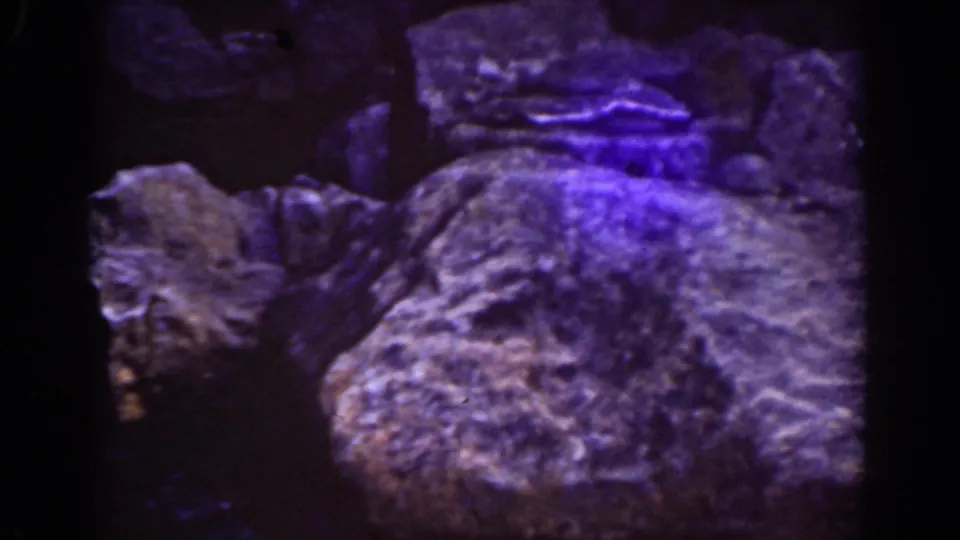 Large foreground boulder and dark stream terrain in 1939 8mm Mount Washington footage, tinted by blue-purple film aging and low exposure.