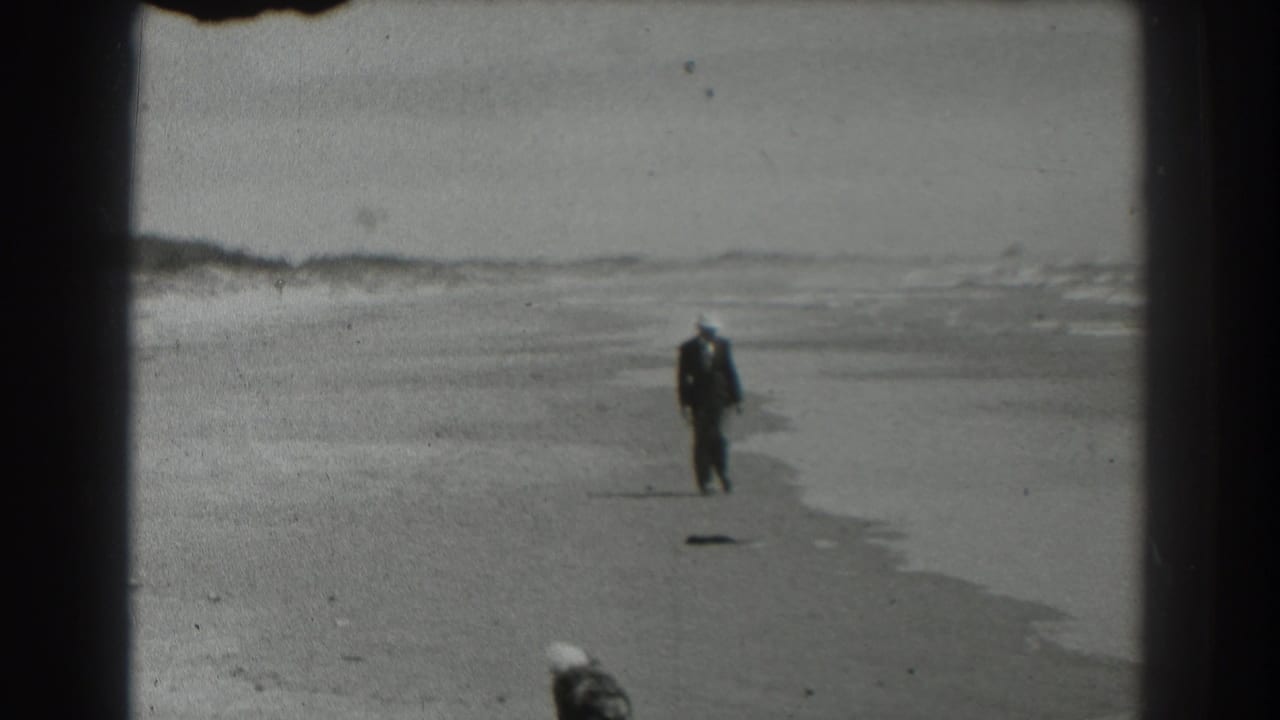 Man in a dark suit and white hat walking on a wide beach near the surf.