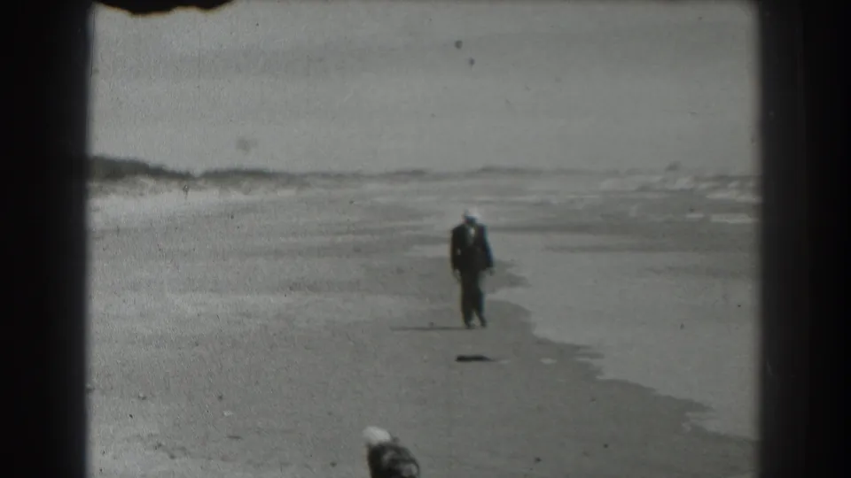 In 1939 San Francisco home-movie footage, a suited man in a white hat walks toward the camera along a broad sandy beach.