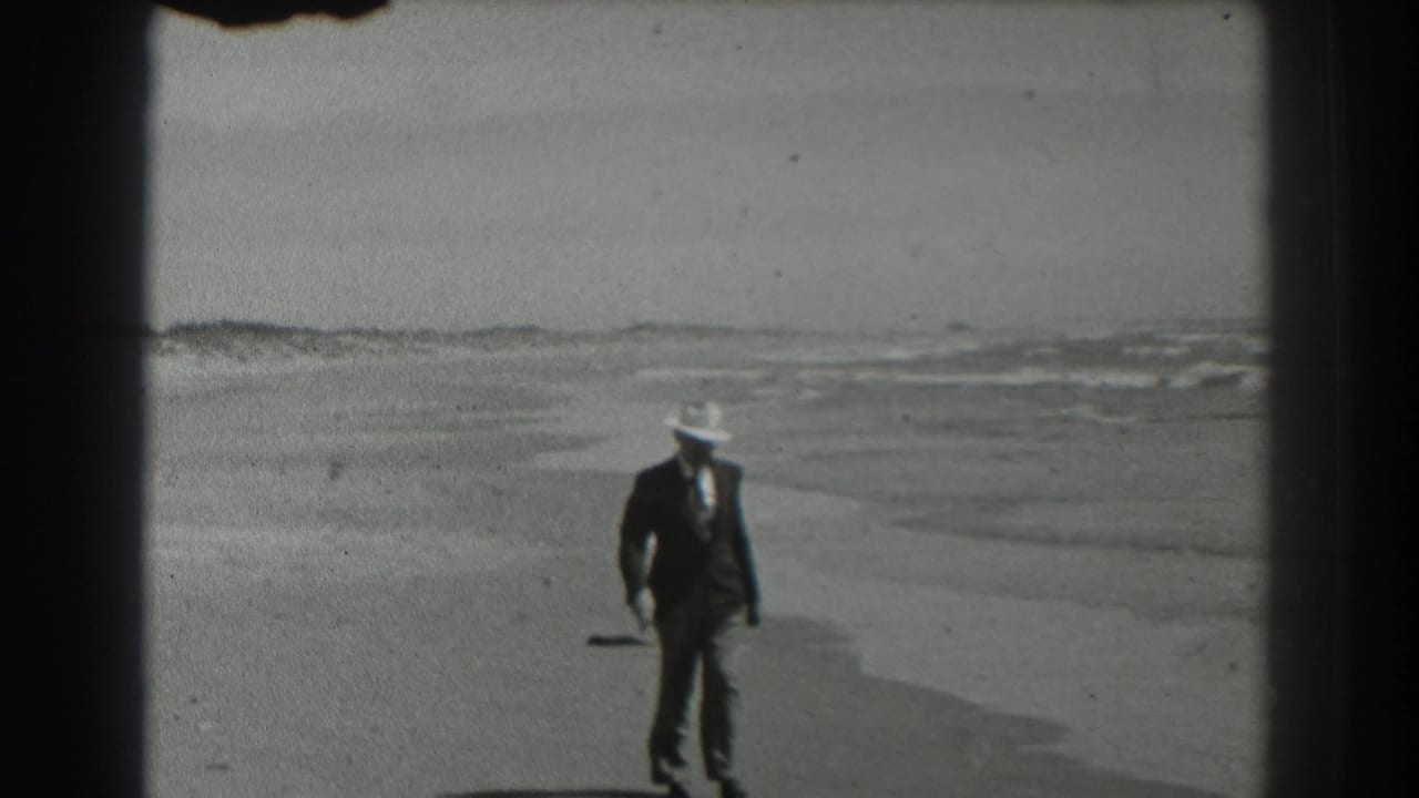 The man in suit, tie, and white hat walks nearer the camera on a San Francisco beach, with low waves behind him.