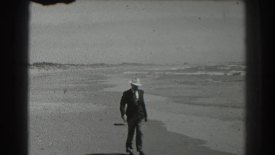 The man in suit, tie, and white hat walks nearer the camera on a San Francisco beach, with low waves behind him.