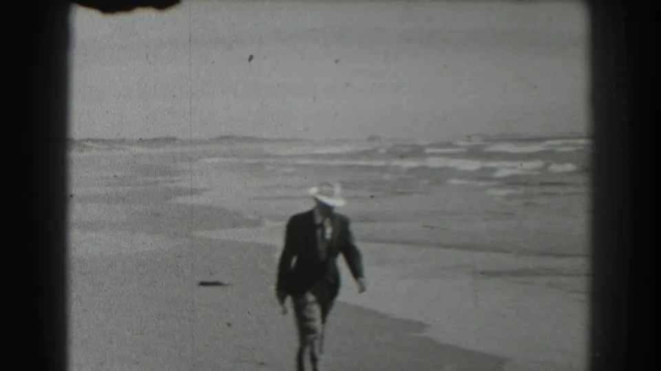 A suited man in a white hat passes closer to the camera on the beach, with the surf and distant shore visible behind him.